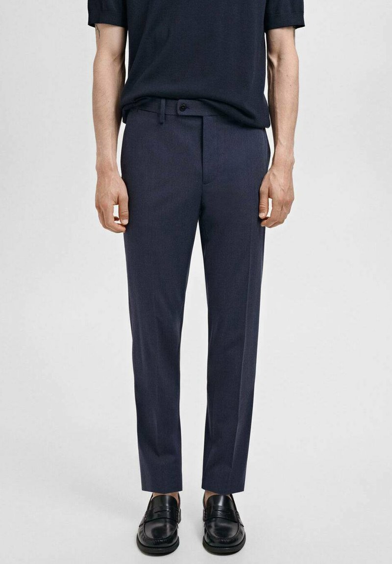 Pantalons taillés en marine en tissu lisse, présentant une coupe ajustée, des passants de ceinture et une fermeture à un bouton. Associés à des chaussures noires.