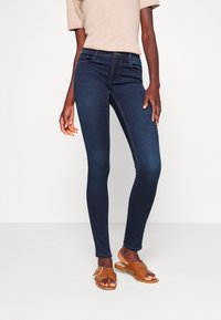 Jean skinny bleu foncé en denim ; avec une taille haute et des coutures contrastantes. Associé avec un haut beige et des sandales marron.