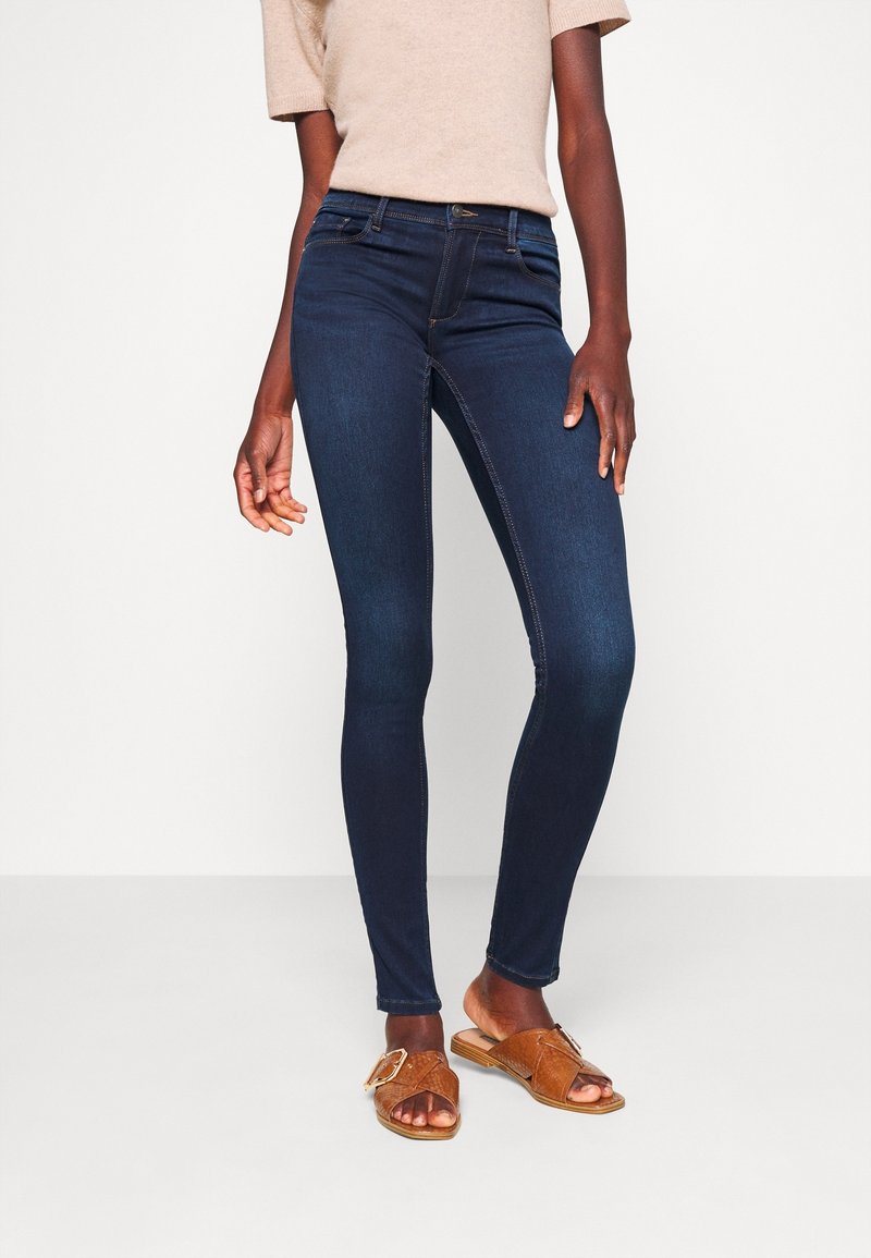 Jean skinny bleu foncé en denim ; avec une taille haute et des coutures contrastantes. Associé avec un haut beige et des sandales marron.