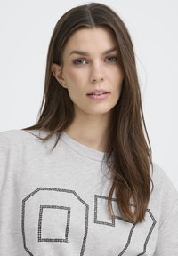 Gaiši pelēka sporta džempera ar melniem rhinestone akcentiem ciparu "07" formā, apaļu kakla izgriezumu un brīvu piegriezumu.