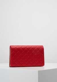 Punainen tilkkukuvioinen tekonahkainen lompakko, jossa on sileä pinta ja ketjuviimeistely. Lompakko on suorakulmainen ja siinä on läppäkiinnitys.