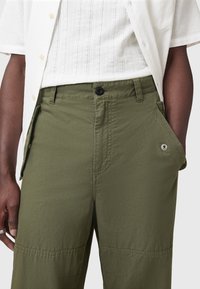Pantalons cargo vert olive avec poche zippée, fermeture à bouton et tissu texturé. Conçus avec des coutures visibles et des poches latérales.
