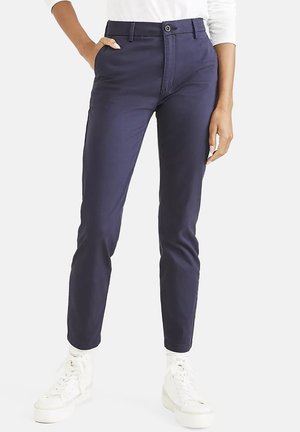 Personne portant un pantalon bleu marine fuselé avec poches avant, un haut blanc à manches longues et des baskets blanches montantes sur fond uni.