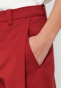 Czerwone spodnie na miarę z płaskim przodem, kieszeniami po bokach i gładką fakturą, z talią wyposażoną w szlufkę na pasek.