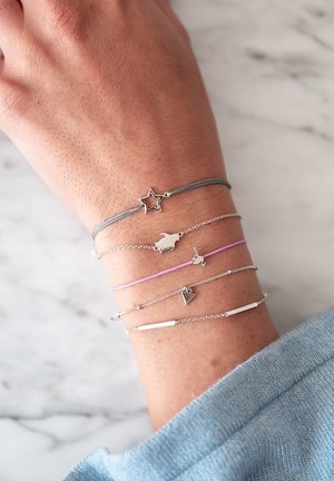 Fem armbånd på et håndled: et gråt snor med en sølvstjerne, sølvkæder med små dyrecharms, og et sølvstangsarmbånd.