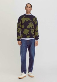 Navy Pullover mit grünem Blattform-Muster, Rundhalsausschnitt und gerippten Bündchen, kombiniert mit Jeans und weißen Sneakers mit Holzsohlen.