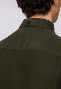 Camicia di corduroy verde oliva scuro caratterizzata da una superficie testurizzata, un colletto classico e un orlo arrotondato. Include un piccolo etichetta nera sul colletto.