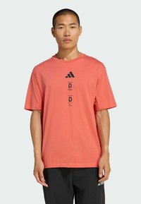 Camiseta de algodón coral de manga corta con cuello redondo. Presenta el logo negro de Adidas y gráficos del equipo Olímpico/Paralímpico impresos en el frente.