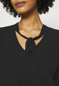 Svart blus med v-ringning och knytinredetail vid kragen, samt två små framfickor. Slät textur och mjukt tyg.