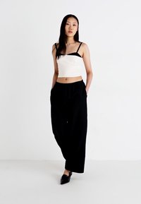 Hvit crop topp med svarte stropper, sammen med svarte vide bukser. Modellen står på en hvit bakgrunn, iført svarte slip-on sko.