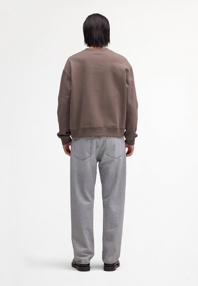Personne aux cheveux noirs vue de dos, portant un sweat-shirt marron, un jean gris clair et des chaussures noires sur un fond blanc.