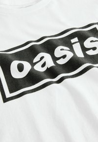 Balta kokvilnas T-kreka ar izteiksmīgu melnu "oasis" logotipu taisnstūra kontūrā. Audums izskatās gluds ar nedaudz spīdumu.