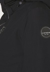 Veste noire avec une poche zippée et un écusson rond avec le logo « ICEPEAK EST. 1996 » sur la zone de l'épaule gauche.