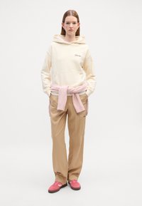 Hoodie beige avec une poche avant, pull rose clair noué autour de la taille, pantalon beige et baskets roses. Tissu lisse, coupe décontractée.
