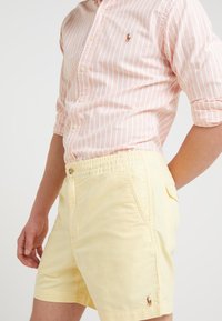 Pánské žluté šortky se bočními kapsami, elastickým pasem a malým vyšitým logem. V kombinaci s růžovou a bílou pruhovanou košilí na knoflík.