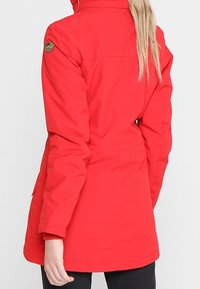 Chaqueta impermeable roja con un cuello alto, cintura ajustable y parche de la marca en el hombro. Tiene mangas largas y una textura suave.