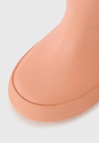Őszibarack színű gumicsizma sima felülettel, lekerekített orral és tapadós, textúrált talppal. Alacsony profillal és varrat nélküli dizájnnal rendelkezik.