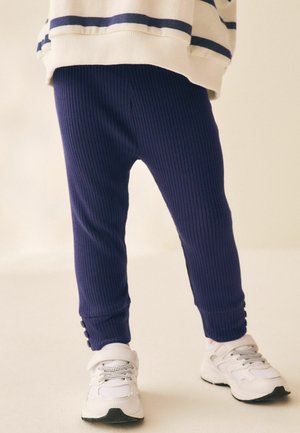 Bambino che indossa pantaloni a coste blu navy, sneaker bianche e una maglia crema con strisce blu scuro, in piedi su uno sfondo chiaro.
