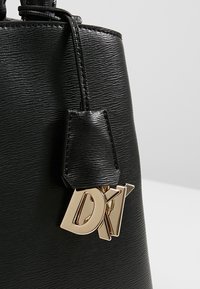 Czarna torebka o fakturze, z pozłacaną metką z logo. Powierzchnia ma subtelny wzór, a torba jest usztywniona w eleganckim stylu.