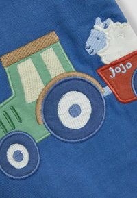 Tissu bleu avec un tracteur vert brodé aux accents marron, de grandes roues et une remorque rouge contenant un mouton blanc avec des détails bleus.