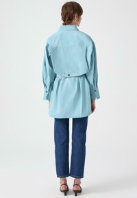Camicia oversize azzurro chiaro con colletto e dettaglio di bottoni sul retro, abbinata a jeans blu scuro a gamba dritta e sandali neri.