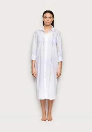 Mujer de pie descalza con un vestido midi blanco de manga larga, abotonado y con un patrón de encaje de ojales, sobre un fondo liso.