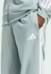 Svijetlozeleni sportski hlače od glatkog materijala, s bijelim trostrukim uzorkom i logotipom, elastičnom padajućom trakom i bočnim džepovima.