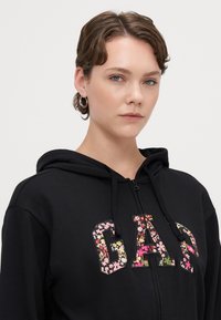 Črna zip-up hoodie s cvetličnim vzorcem na logotipu "GAP" na prsih, z nastavljivo kapuco in mehkim, teksturiranim materialom.