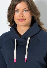 Felpa blu navy con collo a imbuto, dotata di cordini crema con punte rosa. Il materiale sembra morbido, con un design semplice e funzionale.