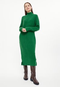 Grünes gestricktes Rollkragenkleid mit gerippter Textur, langen Ärmeln und seitlichen Schlitzern am Saum, kombiniert mit braunen Absatzstiefeln.