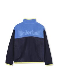 Fleecejakke med en blå øverste del og mørk marineblå nedre del. Har gule detaljer langs kragen og kanten. Timberland-logo vist.