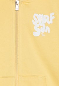Sudadera amarilla con cremallera y gráfico en blanco con las palabras "SURF SUN". Fabricada en tejido suave, con cremallera en la parte delantera y dos bolsillos.