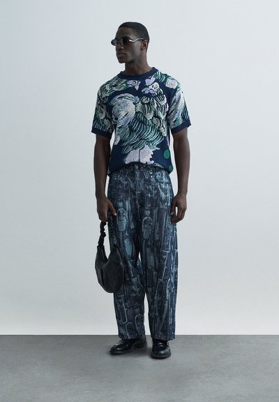 Πλεκτό πουκάμισο σε ναυτικό μπλε με πράσινες λεπτομέρειες, συνδυασμένο με τζιν παντελόνια φαρδιάς γραμμής με περίτεχνο τύπωμα. Μαύρα δερμάτινα παπούτσια και τσάντα.