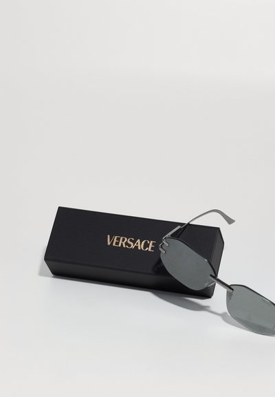 Saulesbrilles ar pelēkiem stikliem un metāla rāmjiem, atpūšoties uz melnas taisnstūra formas kastes, kurā ir zelta "VERSACE" logo.