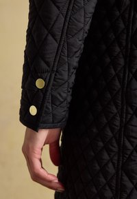 Main reposant près du poignet d'une manche de veste matelassée noire avec deux boutons-pression dorés sur un fond beige.