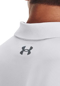 Hvid polo shirt med et tekstureret stof, med et gråt Under Armour-logo på bagkraven. Klassisk kravedesign og minimal detaljer.