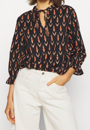 Femme portant un chemisier noir à manches longues avec un motif géométrique orange et un col à nouer avec pompons, associé à un pantalon taille haute blanc cassé.