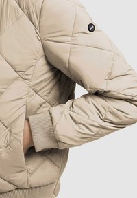 Beżowa pikowana kurtka bomber z prążkowanymi mankietami i obszyciem, z czarnym logo na rękawie, prezentująca gładkie, teksturowane wykończenie.