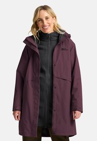 Langer lila wasserdichter Mantel mit Kapuze, Fronttaschen und Druckknopfverschlüssen, über einem schwarzen Fleece-Oberteil getragen. Glatte Textur, lässige Passform.