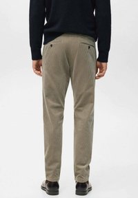 Man draagt beige ribfluwelen broek met achterzakken en donkerbruine schoenen, staand tegen een effen lichte achtergrond.