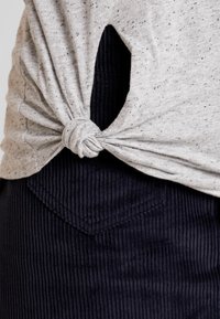 Heathergrå prickig skjorta knuten ovanför midjan över svarta kordflöjlbyxor med en synlig bakficka.