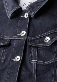 Mörkblå denimjacka med silverfärgade tryckknappar, spetsig krage och kontrasterande vit sömnad, samt två bröstfickor.