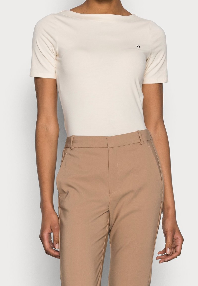 Femme portant un haut court ajusté couleur ivoire et un pantalon beige taille haute, debout devant un fond uni.