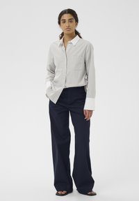Jeune femme portant une chemise gris clair avec des poignets blancs et un pantalon large marine, debout avec une main dans la poche sur un fond blanc.