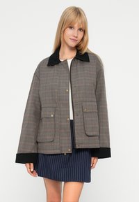 Vrouw in een bruine geruite jas met zwarte kraag en manchetten over een witte top, gecombineerd met een marineblauwe gestreepte rok.