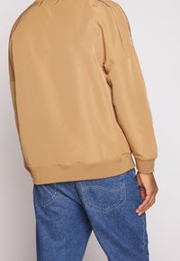 Chaqueta bomber marrón con puños y cintura elásticos, hecha de una tela suave. Combinada con jeans de mezclilla azul claro.
