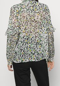 En person visar baksidan av en genomskinlig blus med blommönster och volangärmar, kombinerad med svarta byxor. Tyget har flerfärgade prickar.