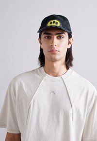 Cappello nero con design grafico giallo, abbinato a una T-shirt morbida e chiara con un piccolo logo. Texture del tessuto liscia e informale.