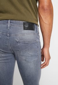 Jeans de mezclilla gris con un diseño ajustado, que presentan un parche de cuero negro en la parte trasera de la cintura y bolsillos traseros cosidos.