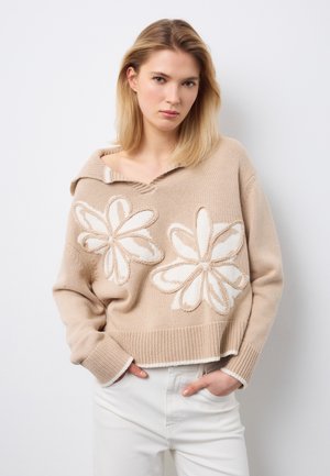 Giovane donna con capelli biondi che indossa un maglione beige con grandi motivi floreali bianchi e pantaloni bianchi, mani nelle tasche, sfondo neutro.